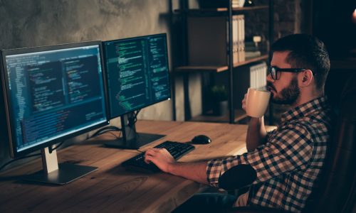 Close up side profile photo handsome mug hot beverage he him his guy coder, typing php css keyboard development outsource IT processing two monitors table office agency wear specs formalwear shirt